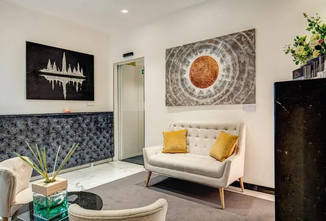 A lobby seating area with a cream sofa, two yellow cushions, an armchair, abstract wall art and a glass-walled lift.