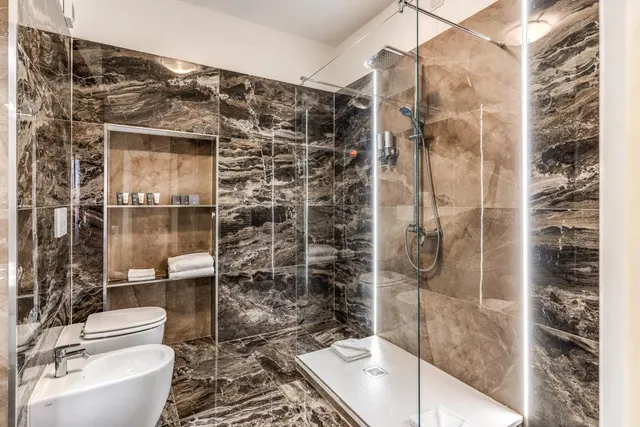 A marble-walled bathroom with a walk-in glass shower, a toilet and a bidet, and towels on a wall niche shelf.