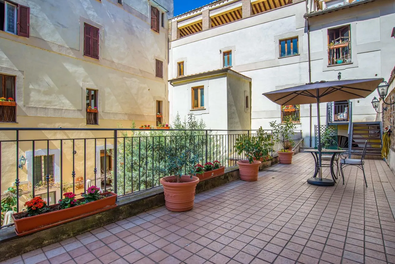 A tiled terrace with potted plants, a table with chairs and a parasol, and a railing overlooking neighbouring buildings.