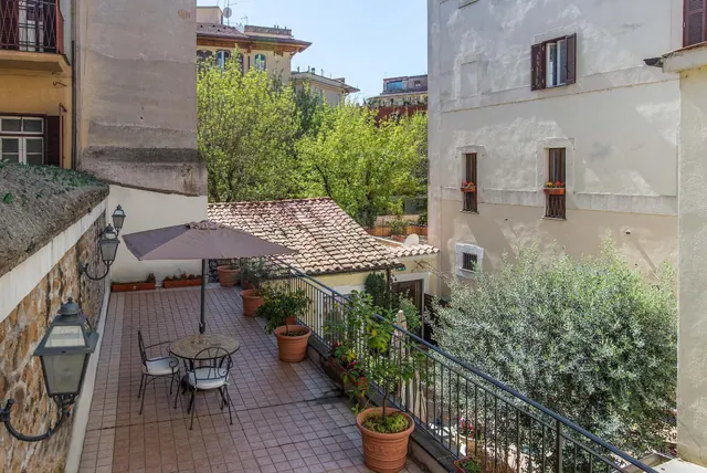 A tiled terrace with a parasol, a metal table and two chairs, potted plants and a railing, overlooking neighbouring buildings and trees.