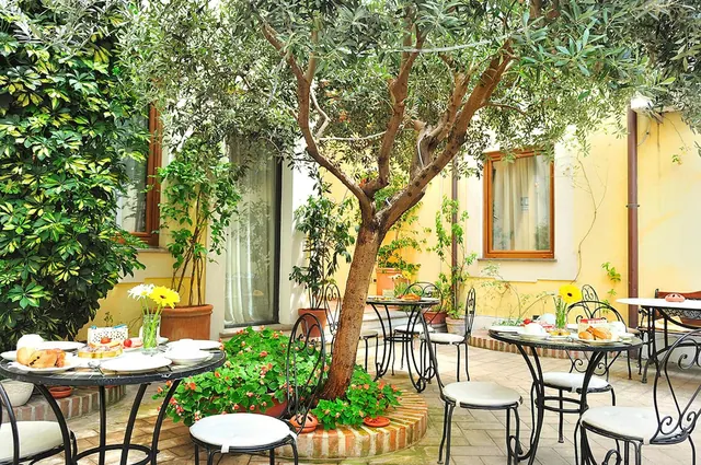 A courtyard with an olive tree, wrought iron tables and chairs with white cushions, some tables set with breakfast food, and yellow walls behind.