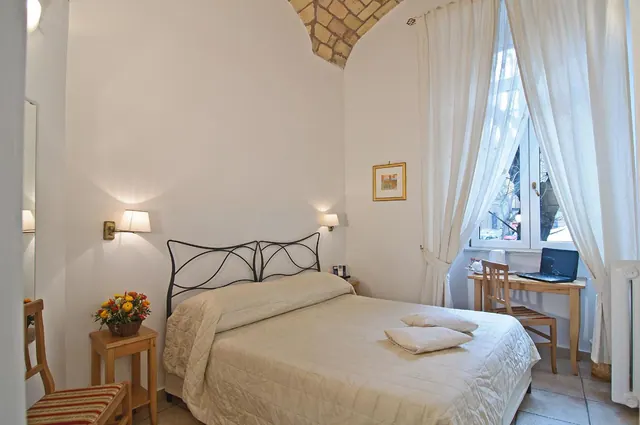 A bedroom with a double bed under a brick vaulted ceiling, a desk with a laptop by the window, and a bedside table with a flower basket.