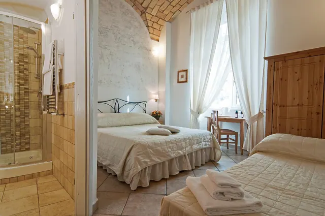 A bedroom with a double bed with a wrought iron headboard, a wooden wardrobe, and a small desk by the window, with a glass-enclosed shower visible through a doorway on the left.