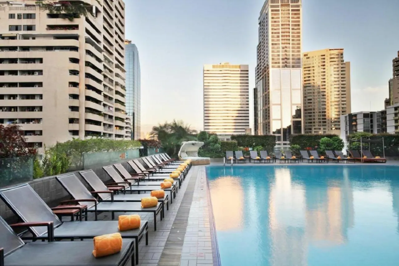 Outdoor pool with rows of sun loungers and orange rolled towels, with high-rise towers rising behind.