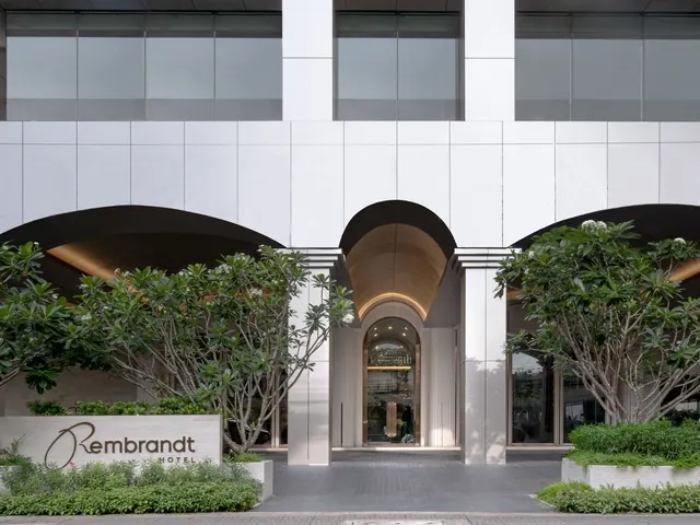 The front facade of the Rembrandt Hotel with an arched entrance, frangipani trees and a stone name sign at the left.