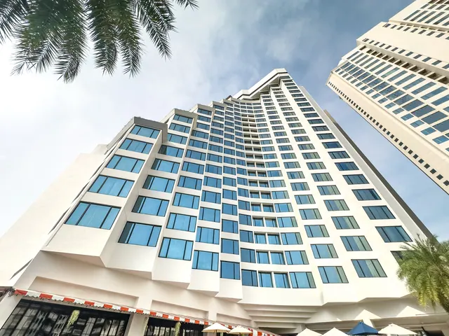 A low-angle view of a white high-rise hotel with rows of blue glass windows, palm fronds at the top left, a second tower on the right, and umbrellas at the base.