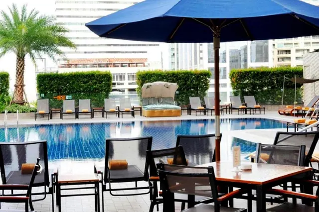 A pool with a blue umbrella, sun loungers and outdoor dining tables, with high-rise buildings and a palm tree behind.