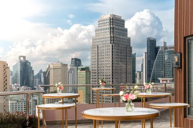A rooftop terrace with round marble-topped tables and flower vases overlooks a city skyline of tall office towers under a partly cloudy sky.