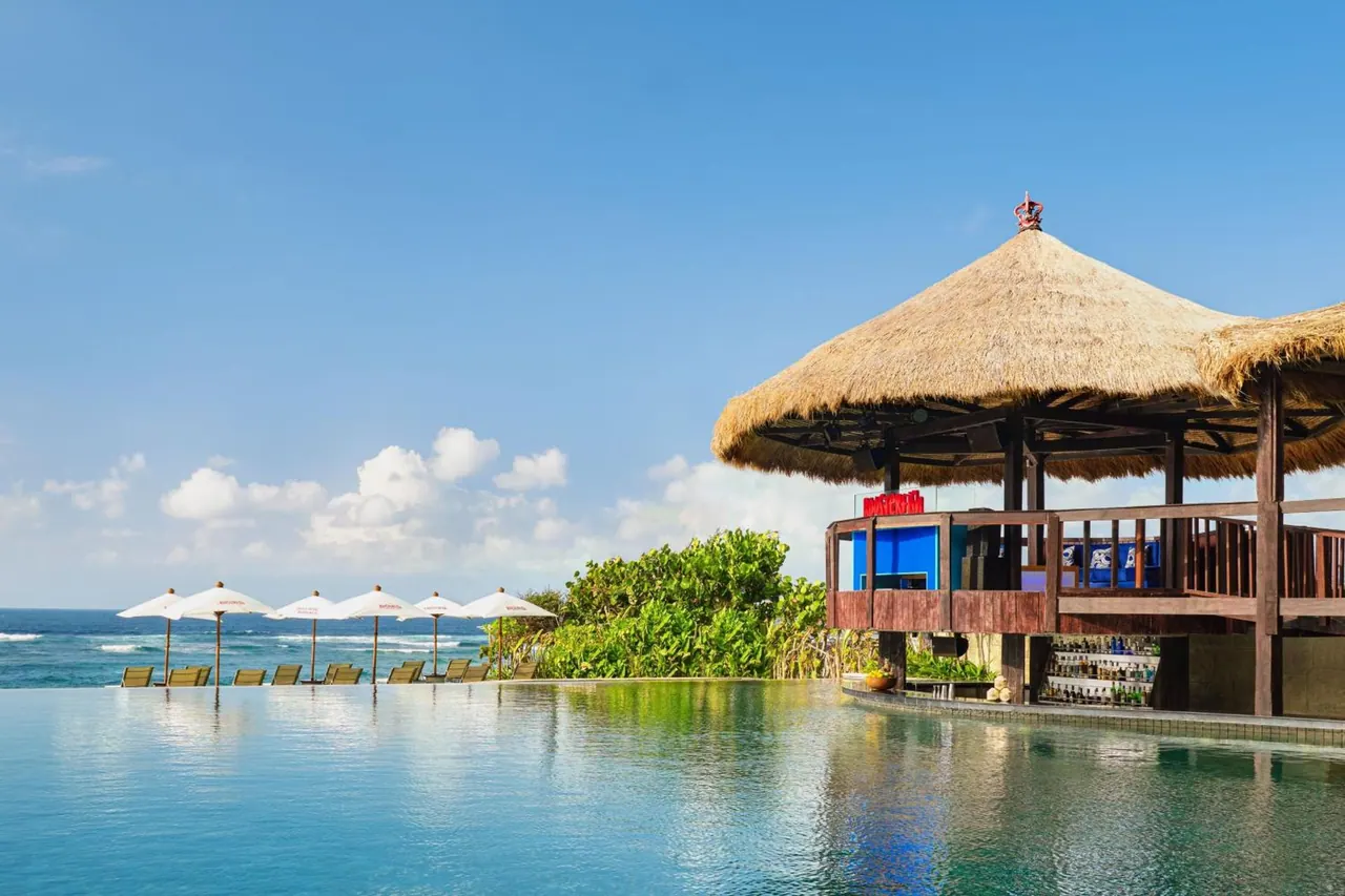 An infinity-edge pool with white parasol umbrellas and sun loungers beside a thatched pool bar, with the sea beyond.
