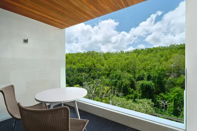 A private balcony with a round white table and two wicker chairs looks out over dense green forest under a partly cloudy sky.