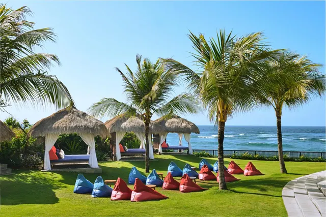 A landscaped lawn with palm trees, thatched daybed cabanas, red and blue bean bags, and the ocean beyond a low fence.