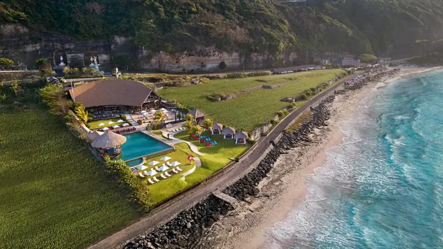 Aerial view of a beachfront resort with a pool, a thatched-roof restaurant, sun loungers and umbrellas, lawns, and the ocean along a rocky shoreline.