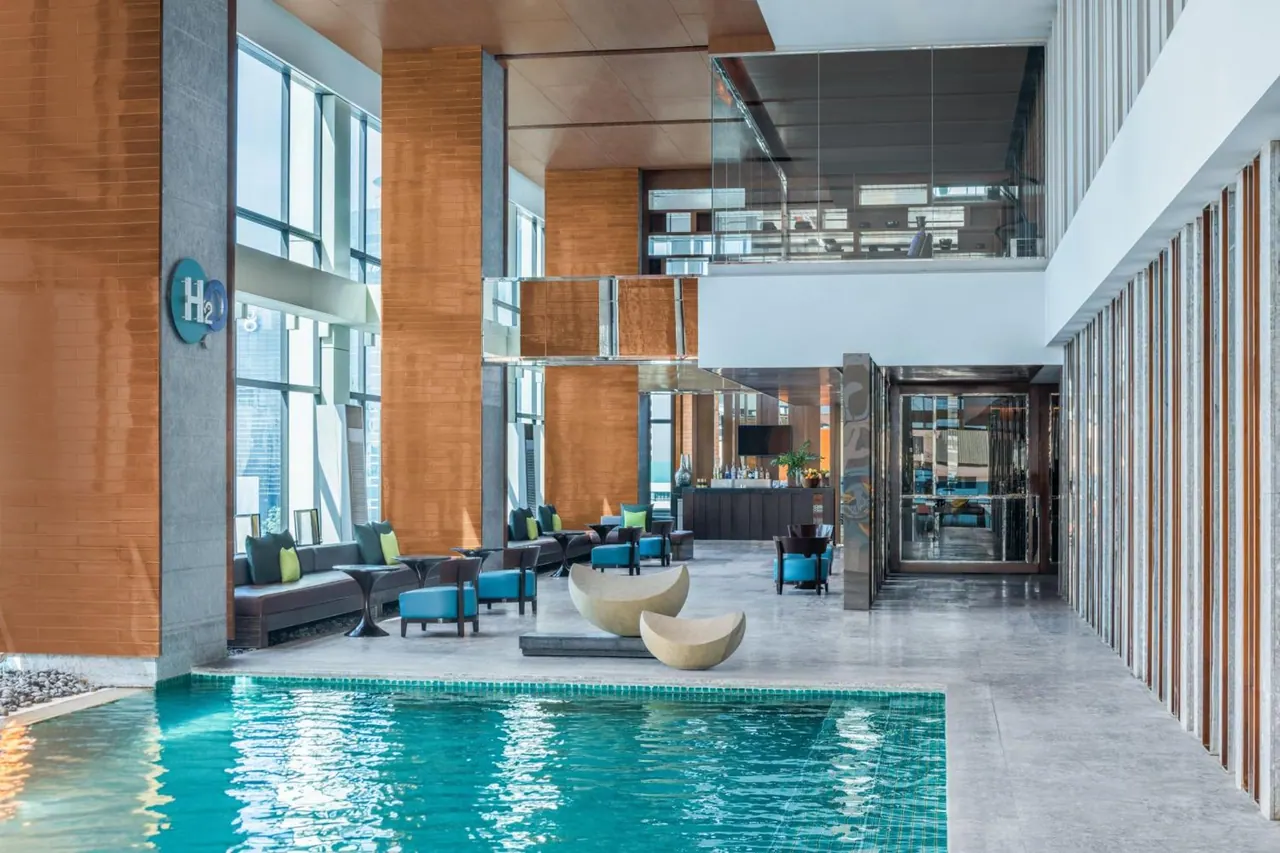 An indoor pool with turquoise water sits beside a lounge area with sofas, armchairs and stone tables under tall windows.