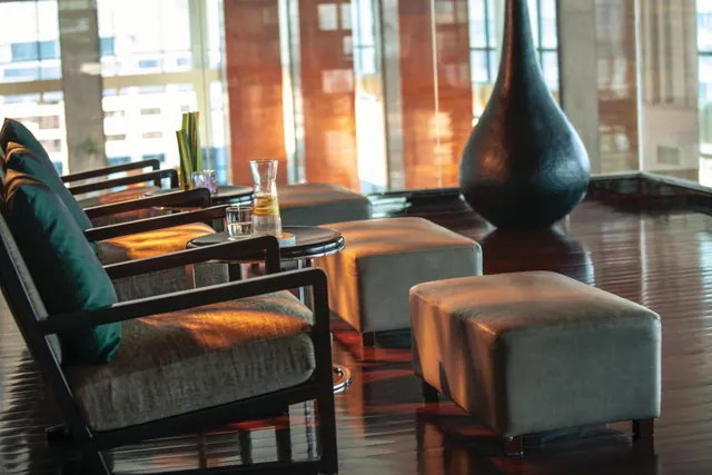 A lounge seating area with grey armchairs and ottomans, side tables holding a water carafe and glasses, a tall dark vase, and large windows over a polished wood floor.