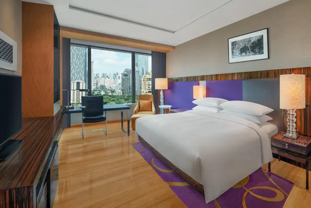 A hotel bedroom with a king bed, a desk and armchair, and floor-to-ceiling windows overlooking a city skyline.