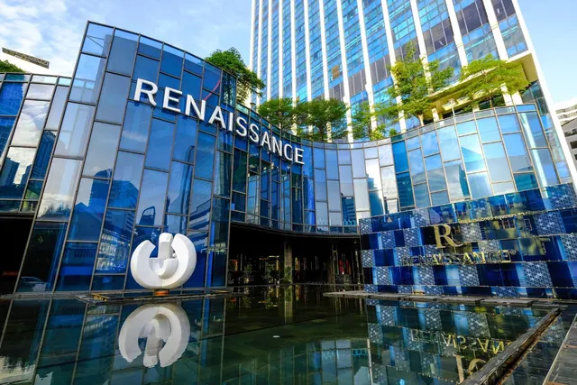 The blue glass facade of the Renaissance building with its name sign, a white abstract sculpture and a reflecting pool in front, with a tall tower behind.