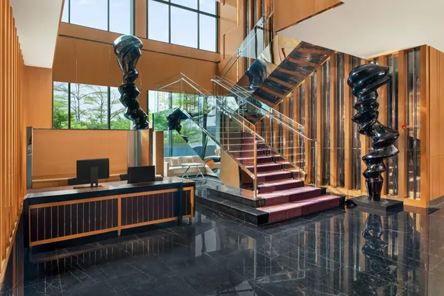 A hotel lobby with a reception desk, a staircase with burgundy carpeted steps, two black sculptures, and tall windows showing trees outside.