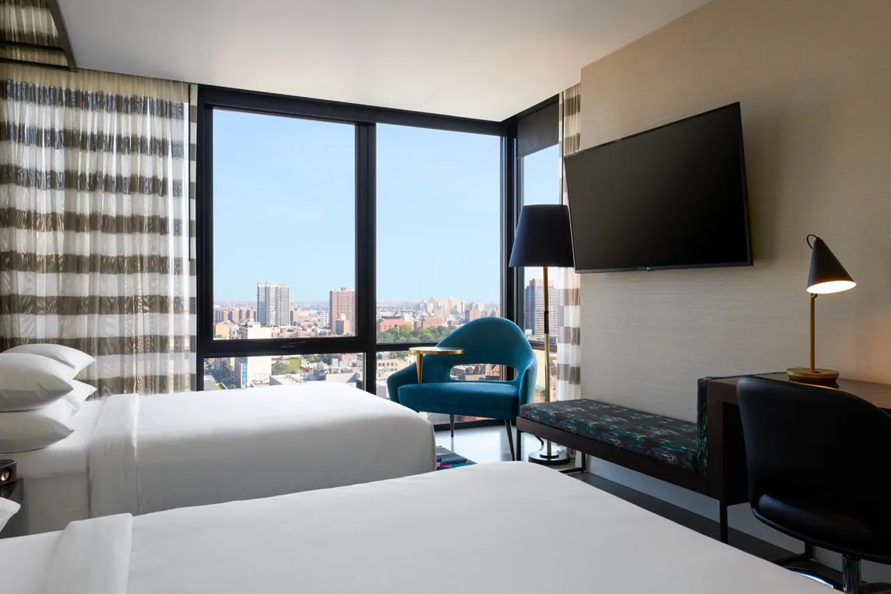 A hotel bedroom with two beds, a blue armchair, a wall-mounted television and a desk, with large windows showing a city skyline.