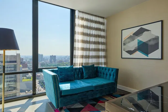 A teal velvet sofa sits beside floor-to-ceiling windows that look out over a city skyline.