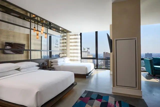 A hotel bedroom with two beds, a wall-mounted television, a teal armchair and floor-to-ceiling windows looking out over a city.
