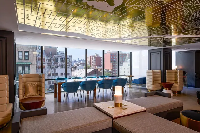 A lounge with a gold mirrored ceiling printed with a street map, cream armchairs, a long dining table with blue chairs, and a view of brick buildings through floor-to-ceiling windows.
