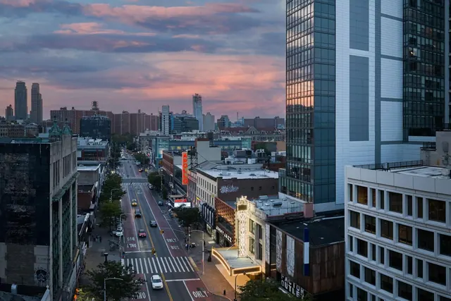 A dusk view from an upper floor down a city street, with tall buildings, a distant skyline under a pink sky, and traffic on the road below.