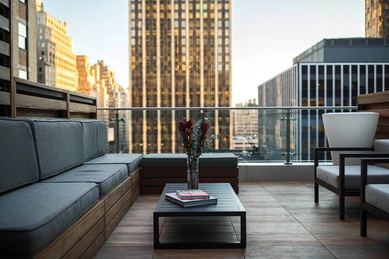 A furnished rooftop terrace with a built-in grey sofa, a dark coffee table holding a vase of flowers and books, and a glass railing overlooking tall city buildings.