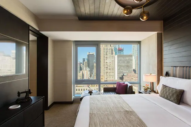 A king bed with white linens faces a large window overlooking high-rise buildings, with a seating area and a dark wood dresser in the room.