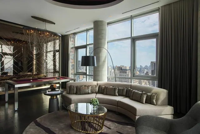 A living area with a curved sofa, floor-to-ceiling windows, a pool table and a view over city buildings.