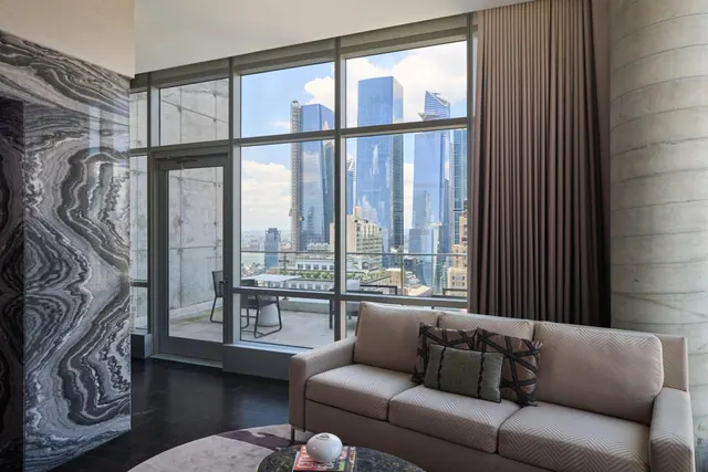 A living room with a light grey sofa, a dark marble accent wall, and floor-to-ceiling windows looking out past a balcony onto tall glass skyscrapers.