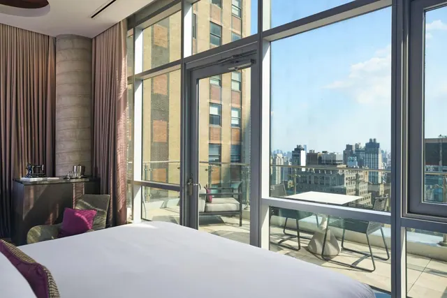A bedroom with a made king bed, an armchair and a sideboard beside floor-to-ceiling windows that open onto a balcony with a table and chairs, with a city skyline in the distance.