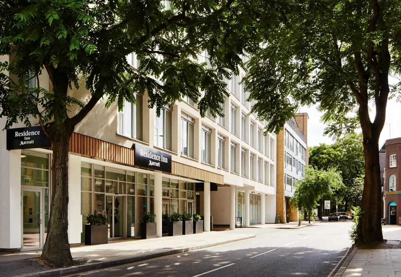The exterior of a Residence Inn by Marriott building is shown with planters at its entrance and trees lining the street.