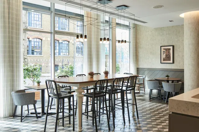 A dining area with tall bar tables and stools, grey armchairs, pendant lights and large windows overlooking a brick building.