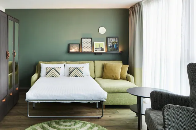 A hotel room with a green sofa bed opened out into a made bed with white linen, beside a green wall with framed prints, a wardrobe and sheer curtains at the window.