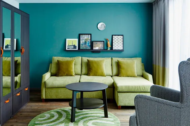A living area with a green sofa and cushions, a round black coffee table on a green rug, a teal wall with framed pictures on a shelf, and a patterned armchair at the right.