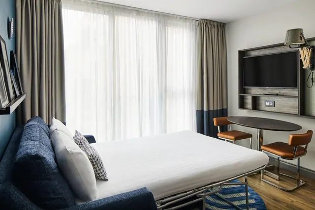 A hotel room with a made-up sofa bed, a wall-mounted television, and a round table with two tan chairs beside a large window with sheer curtains.