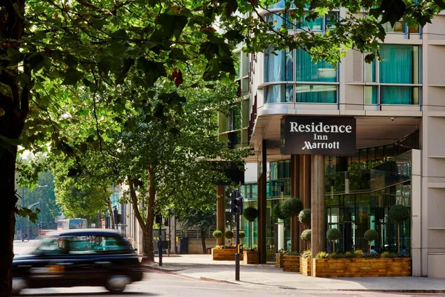 Exterior of the Residence Inn by Marriott building with its entrance sign, planters and tree-lined street, with a blurred black taxi passing in the foreground.