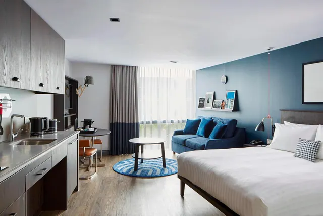 A studio hotel room with a made bed, a blue sofa, a round coffee table, a kitchenette with a sink and kettle, and a bistro table by the window.