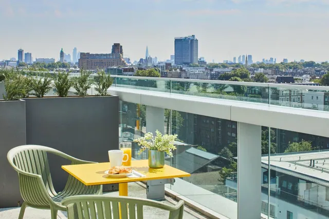 A rooftop terrace with a yellow table set with a croissant, orange juice and a mug, green chairs, and a view over the London skyline.