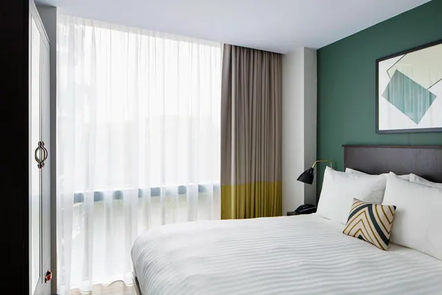 A bedroom with a made king bed, a green accent wall, and sheer curtains over a floor-to-ceiling window.
