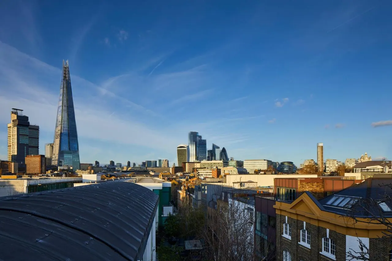 View over rooftops toward the City of London skyline, with The Shard on the left, under a clear blue sky.