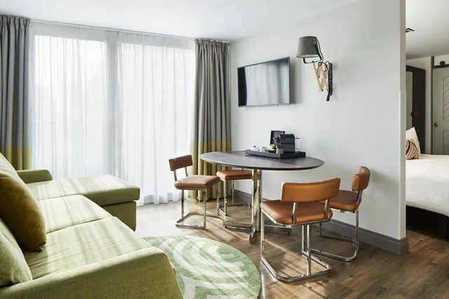 A hotel room with a green sofa, a round dining table with four tan chairs, a wall-mounted television, a coffee machine on a tray, and a bed visible through a doorway.