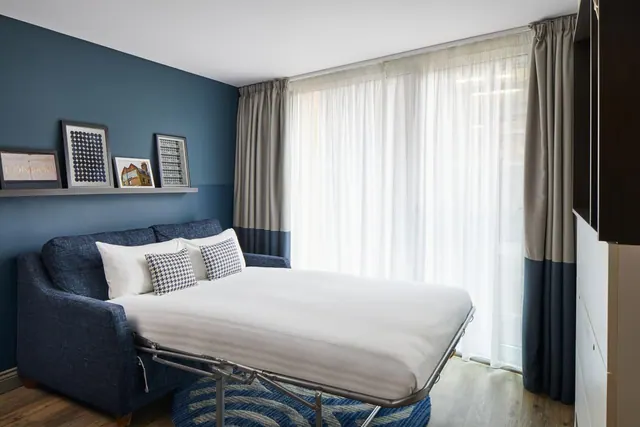 A made-up sofa bed with white bedding and patterned cushions stands in front of sheer-curtained windows, beside a blue accent wall with framed prints.