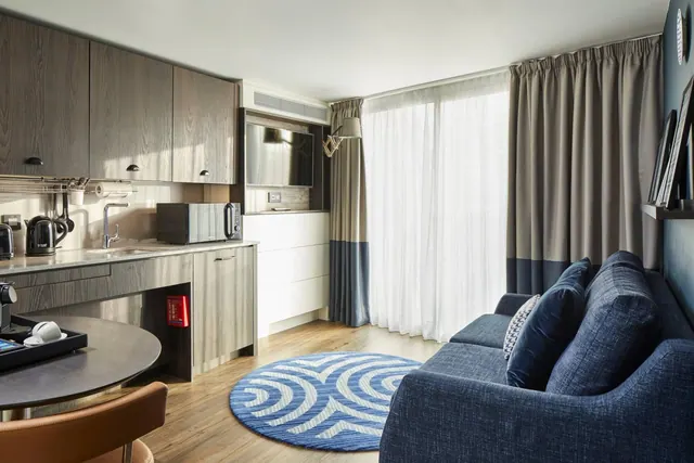A hotel room living area with a kitchenette, round dining table, blue sofa, round blue rug and sheer curtains over a large window.