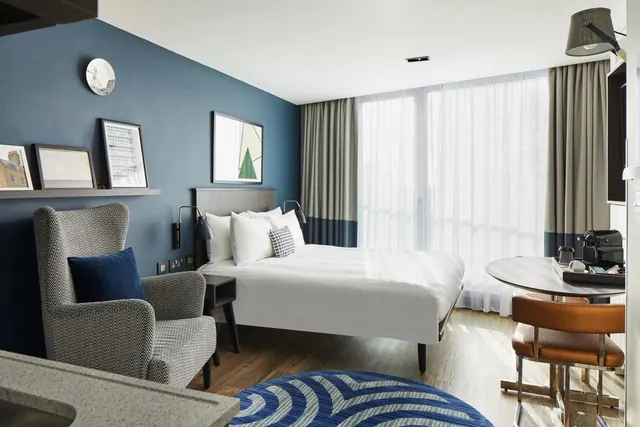 A hotel bedroom with a double bed, a grey armchair beside a blue accent wall, sheer floor-to-ceiling curtains and a round table with a leather chair.