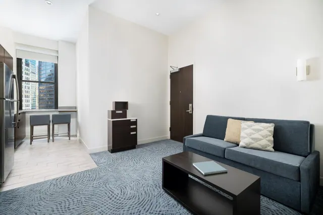 A living area with a blue sofa, a dark coffee table, patterned carpet, a kitchenette with a stainless steel fridge, and a window showing high-rise buildings.