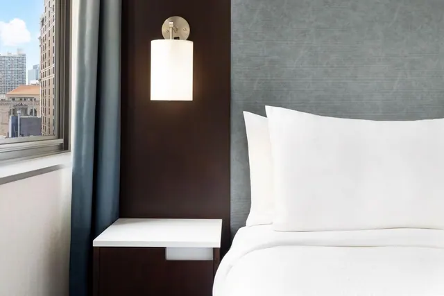 A bedroom corner with a made bed, white pillows, a grey upholstered headboard, a wall lamp above a dark wood nightstand, and a window showing city buildings.