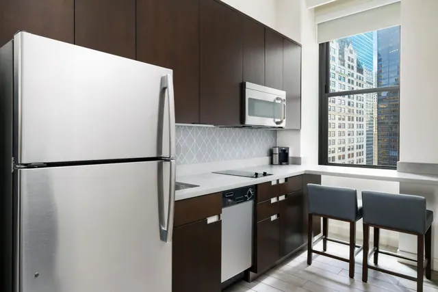 A kitchenette with dark wood cabinets, a stainless steel refrigerator and microwave, two bar stools, and a window showing high-rise buildings.