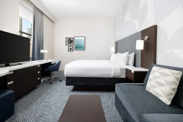A hotel bedroom with a king bed, a work desk and chair, a sofa, a wall-mounted television, and a window showing city buildings.