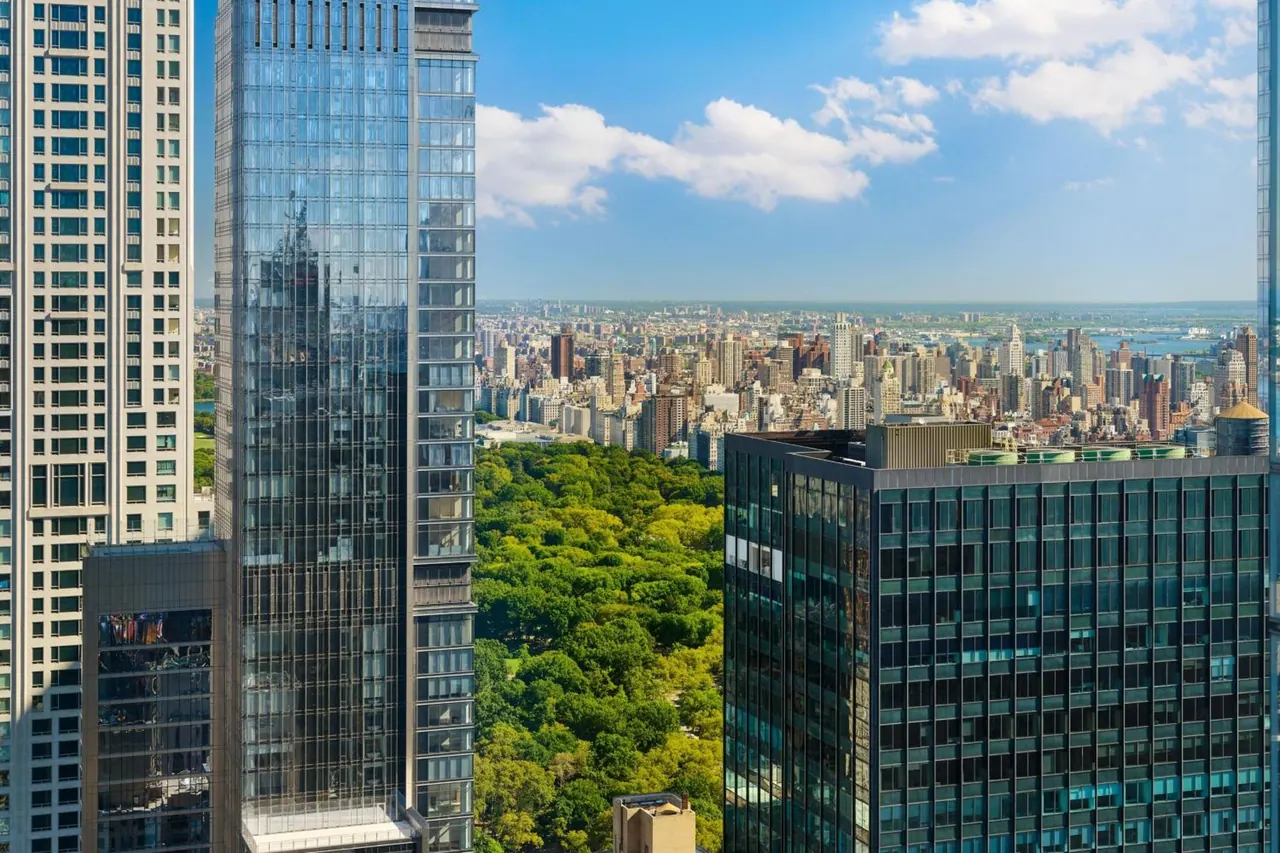 A high-floor view over the green canopy of Central Park and the Manhattan skyline, with glass towers in the foreground under a partly cloudy sky.